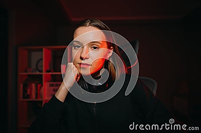 Close-up portrait of an attractive female gamer in a headset around her neck sitting at night in a bedroom with a red light and Stock Photo