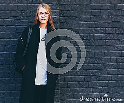 Close-up portrait of a beautiful blonde girl in round fashionable glasses in a black coat and boots near a black brick Stock Photo