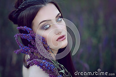 Beautiful blue-eyed lady with perfect make up and plaited hairstyle sitting in the field and holding purple flowers at her face Stock Photo