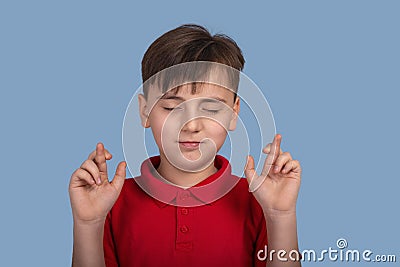 Studio portrait of a of a boy with closed eyes suggesting a desire and showing hands with crossed fingers on blue background in Stock Photo