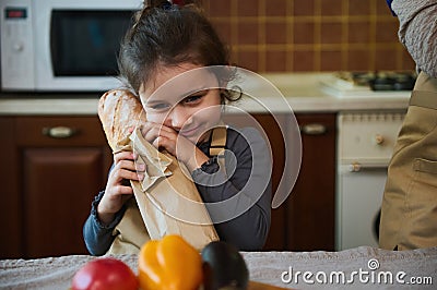 Close-up portrait of a cute little girl hugging a loaf of sourdough bread from family bakery while unpacking grocery bag Stock Photo