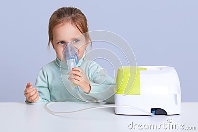 Close up portrait of charming cute small girl uses nebulizer mask for inahlation, has respiratory disease, sits at white desktop, Stock Photo