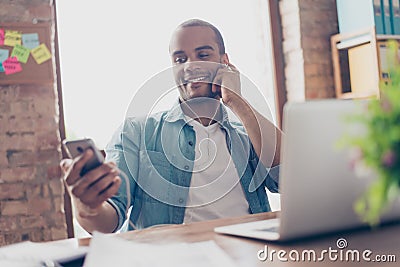 Close up portrait of cheerful young afro guy enjoying listening to nice song at his work place, on his smart phone, wearing casual Stock Photo
