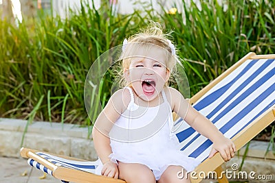 Close up portrait of cute emotional blondy toddler girl in white dress sitting on the deckchair and yelling. City park recreation Stock Photo