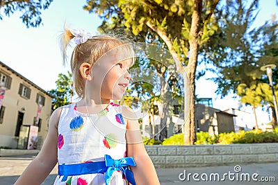 Close up portrait of cute little blondy toddler girl in dress looking aside in the city park with buildings and trees on the backg Stock Photo