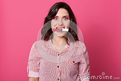 Close up portrait of darkhaired female dresses striped shirt, posing isolated over rose background, charming girl bitting lipstick Stock Photo