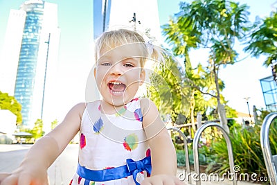 Close up portrait of emotional cute little blondy toddler girl in dress in the city park with modern skyscraper buildings and tree Stock Photo