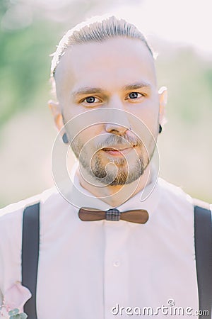 The close-up portrait of the groom with the nose piercing and wooden butterfly in the green field. Stock Photo