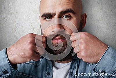 A close-up portrait of handsome balded man having thick black eyebrows, beard and moustasche, dark eyes wearing casual jean shirt Stock Photo