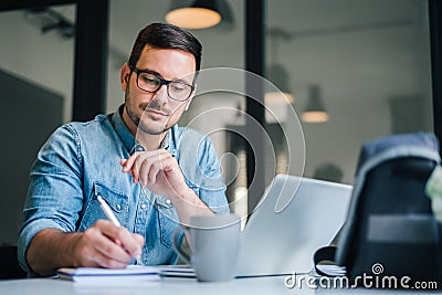 Close up portrait of handsome man working from home office taking reading and writing notes in note pad while working on laptop Stock Photo