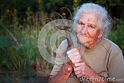 Close-up portrait of an old woman with gray hair holding a rusty pitchfork or chopper in her hands, face in deep wrinkles, Stock Photo