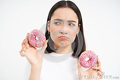 Close up portrait of sad asian woman, upset being on diet, showing two glazed pink doughnuts, tempted to eat junk food Stock Photo