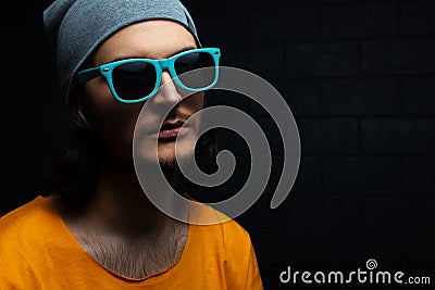 Close-up portrait of serious young man on black background; wearing blue sunglasses, grey hat and orange shirt. Stock Photo