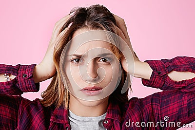 Close up portrait of stressful female model touches head and suffers from headache, looks frusrtated at camera, dressed in checker Stock Photo