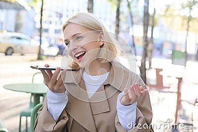 Close up portrait of young smiling woman, lively recording voice message, talking into smartphone microphone, sitting Stock Photo