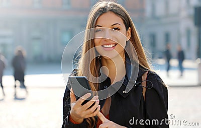 Close up portrait of young woman typing on smart phone looking at camera in city street in spring time Stock Photo