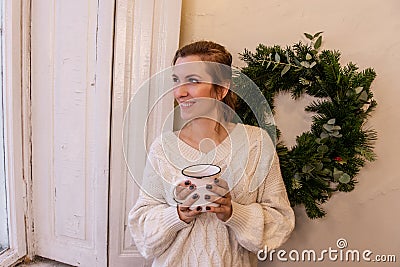 Close-up portraits of young stylish woman with white cup in hands near green Christmas wreath Stock Photo