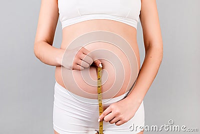 Close up of pregnant woman in white underwear checking her growing belly with a tape measure at gray background. Inch measurement Stock Photo