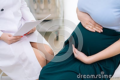 Close-up of pregnant woman holding hands on belly while sitting in tight medicine office. Stock Photo