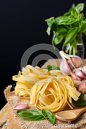 Close-up of raw tagliatelle nests on burlap with wooden spoon, basil and garlic, selective focus, on rustic table and black backgr Stock Photo