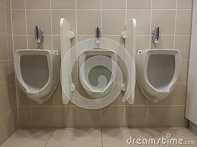 Close up of a row of three white ceramic urinals fixed on a beige tile wall separated by privacy walls Stock Photo