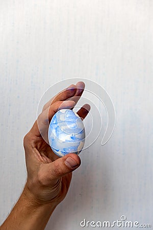 selective focus male hand stained with blue paint holding a freshly painted Easter egg against a white wall Stock Photo