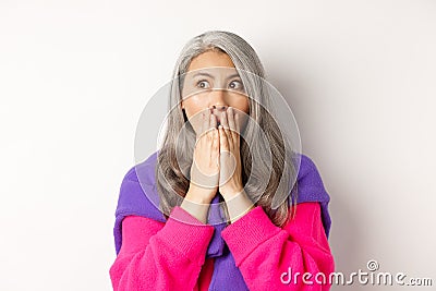 Close-up of shocked asian middle-aged woman with grey hair, gasping and covering mouth startled, looking left, realising Stock Photo