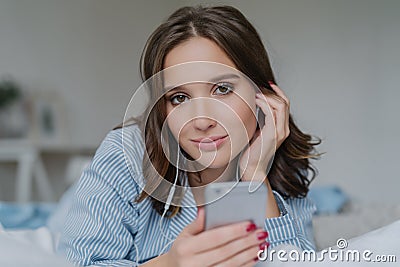 Close up shot of attractive brunette young woman downloads songs from network, uses smart phone for entertainment, dressed in nigh Stock Photo