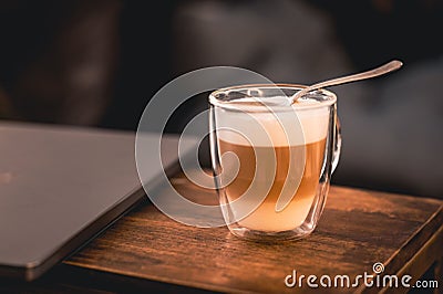 Close-up shot of a cup of Latte coffee with layers and frothy milk on a wood table with laptop Stock Photo