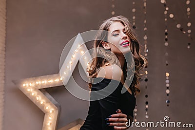 Close-up shot of flirtatious lady in elegant dark outfit posing in gray studio with star decoration. Stock Photo