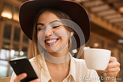 Close up of a smiling lovely girl in hat sitting at the cafe table indoors, holding cup of tea, Stock Photo