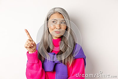 Close-up of stylish korean senior woman with grey hair looking and pointing upper left corner, showing promotion offer Stock Photo