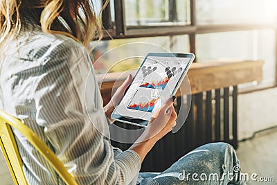 Close-up of tablet computer with charts, graphs, diagrams on screen in hands of young woman sitting in room on chair. Stock Photo