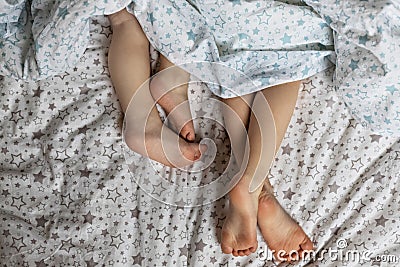 Close-up of two pairs of toddler girls feet on the bed under the blanket. Light blue and beige tones Stock Photo