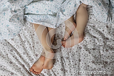 Close-up of two pairs of toddler girls feet on the bed under the blanket. Light blue and beige tones Stock Photo