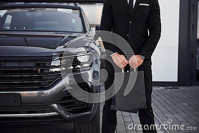 Close up view of businessman in black suit and tie stands near modern automobile with shopping bags in hands Stock Photo