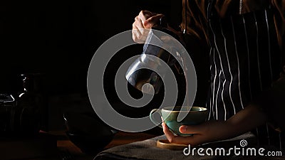 Female barista spills hot coffee from coffee pot to cup in dark background Stock Photo