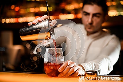 Close-up view of hand of bartender holds shaker with strainer and pours steaming drink into glass. Stock Photo