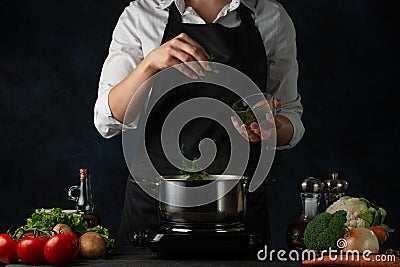 Close-up view of the professional chef in black apron adding chopped parsley in boiling water for soup on dark blue background. Stock Photo