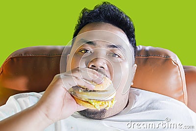 Voracious obese man eating a burger on the sofa Stock Photo