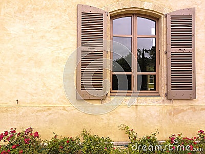 Close-up on a window and its light brown shutters, ocher wall and alley of small red roses in bloom, against the Stock Photo