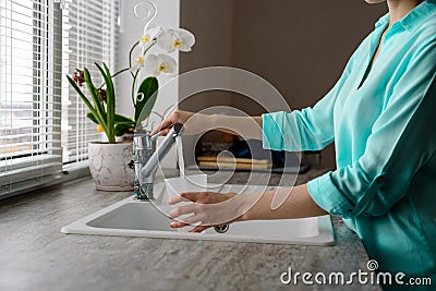 Close-up of a woman collects water in a plastic glass from the tap in the kitchen sink in front of the window Stock Photo
