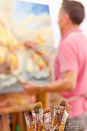 Close up of wooden flask with paint brushes, with blurred man painting as background Stock Photo