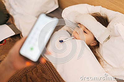 Close-up of worried mother sitting on bed beside sick daughter with high fever and calling on phone to family doctor. Stock Photo