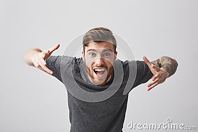 Close up of young cheerful bearded tattooed good-looking guy with stylish hairstyle in casual gray shirt spreading hands Stock Photo