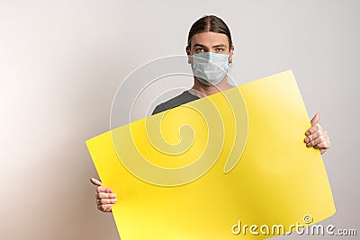 Close up of a young man with protective mask against virus epidemy is holding an empty yellow cardboard against white Stock Photo