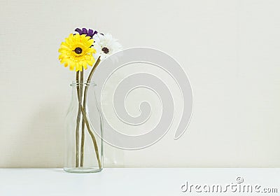 Closeup artificial colorful flower on transparent glass bottle on blurred white wood desk and cream color wallpaper at the wall Stock Photo