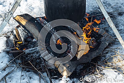 Closeup campfire in the snow and caked pot in soot over the fire on tripod, winter outdoor cooking at the campsite Stock Photo