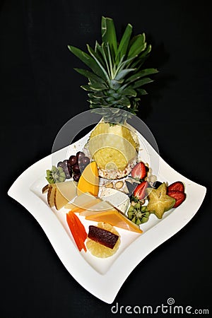 Closeup display of assorted sliced fruits and cheeses with sculpted Pineapple on a white plate with a black background Stock Photo