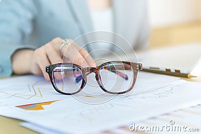 Closeup eyeglasses and female hand on background laptop and graph report data on wood desk working at the office Stock Photo